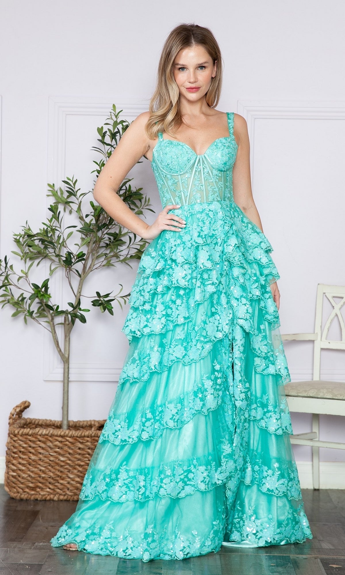 A woman wears the Sheer-Corset Long Ruffled Prom Dress: Poly USA 9410 in turquoise, with a tiered skirt and floral details. She's indoors, framed by a potted plant and white chair that enhance the elegant setting.